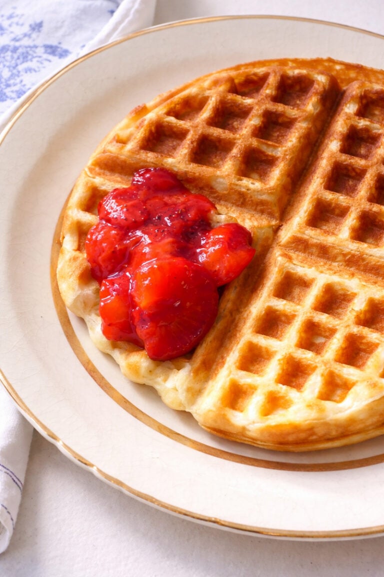 A golden brown yeast waffle on a beige-rimmed plate, topped with a spoonful of bright red strawberry compote, sits on a white tablecloth.