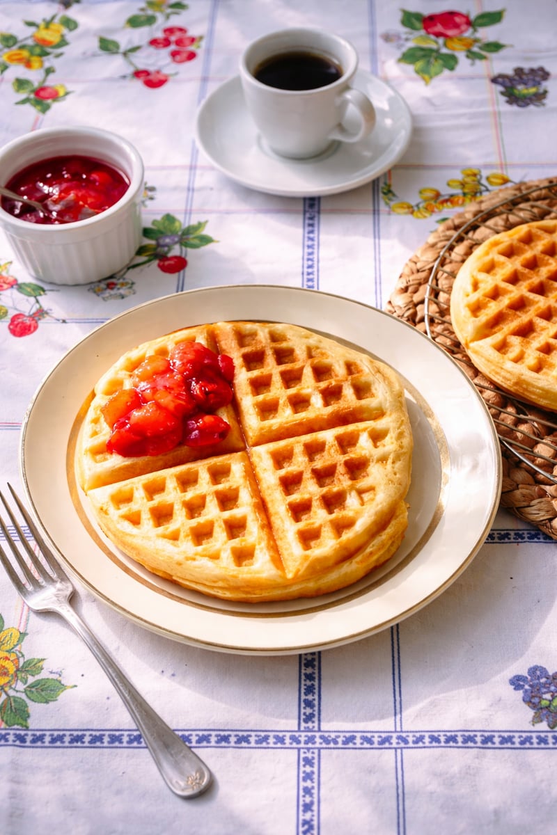 A stack of golden yeast waffles topped with strawberry sauce sits on a plate, fork beside it. In the background are more waffles, a cup of coffee, and a ramekin of strawberry sauce on a floral tablecloth.