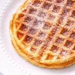A golden brown yeast waffle sprinkled with powdered sugar sits on a white, textured plate.