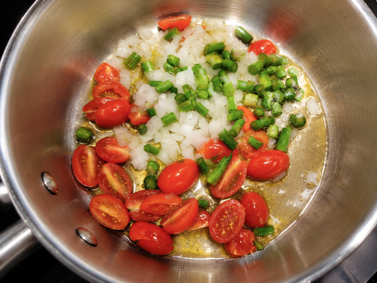 Chopped onions, green peppers, and halved cherry tomatoes sautéing in olive oil inside a stainless steel pan.
