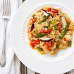 A white plate of vegetable risotto with tomatoes, asparagus, and mushrooms sits on a rustic wooden table, next to a fork and white napkin.