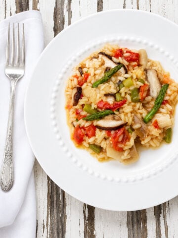 A white plate of vegetable risotto with tomatoes, asparagus, and mushrooms sits on a rustic wooden table, next to a fork and white napkin.