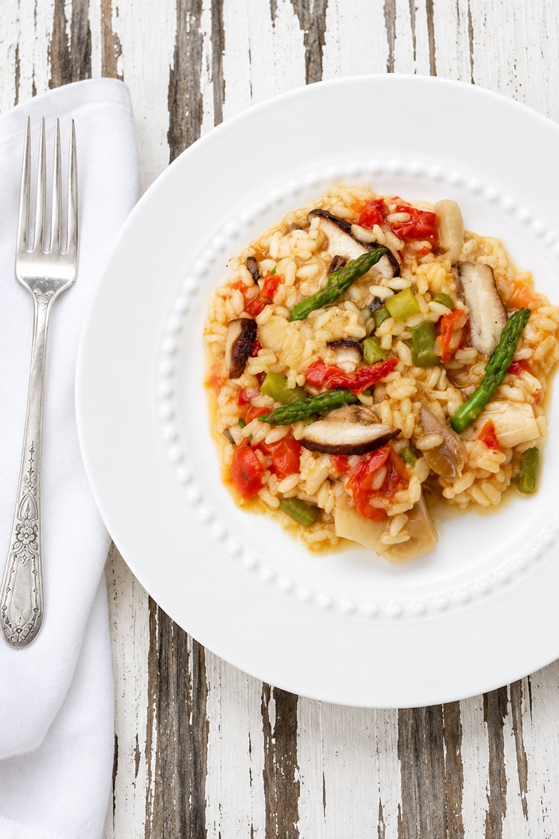 A white plate of vegetable risotto with tomatoes, asparagus, and mushrooms sits on a rustic wooden table, next to a fork and white napkin.