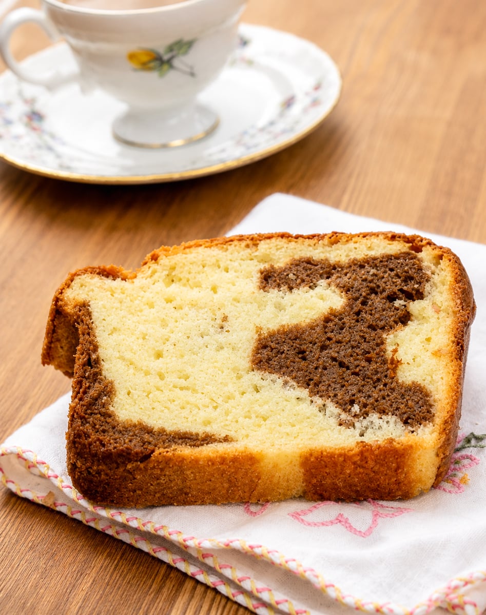 A slice of marble cake with vanilla and chocolate swirls rests on a white napkin, with a floral teacup and saucer in the background on a wooden table.