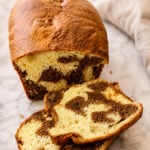 A loaf of marble bread with a golden-brown crust sits on a marble surface. Two slices are cut, revealing a swirled pattern of chocolate and vanilla bread inside. A beige cloth is in the background.