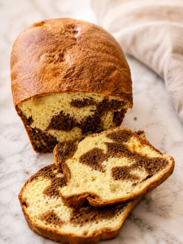 A loaf of marble bread with a golden-brown crust sits on a marble surface. Two slices are cut, revealing a swirled pattern of chocolate and vanilla bread inside. A beige cloth is in the background.