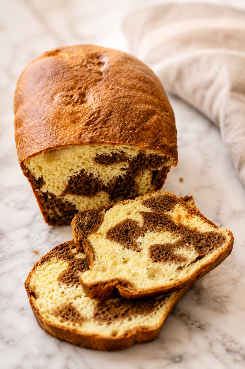 A loaf of marble bread with a golden-brown crust sits on a marble surface. Two slices are cut, revealing a swirled pattern of chocolate and vanilla bread inside. A beige cloth is in the background.