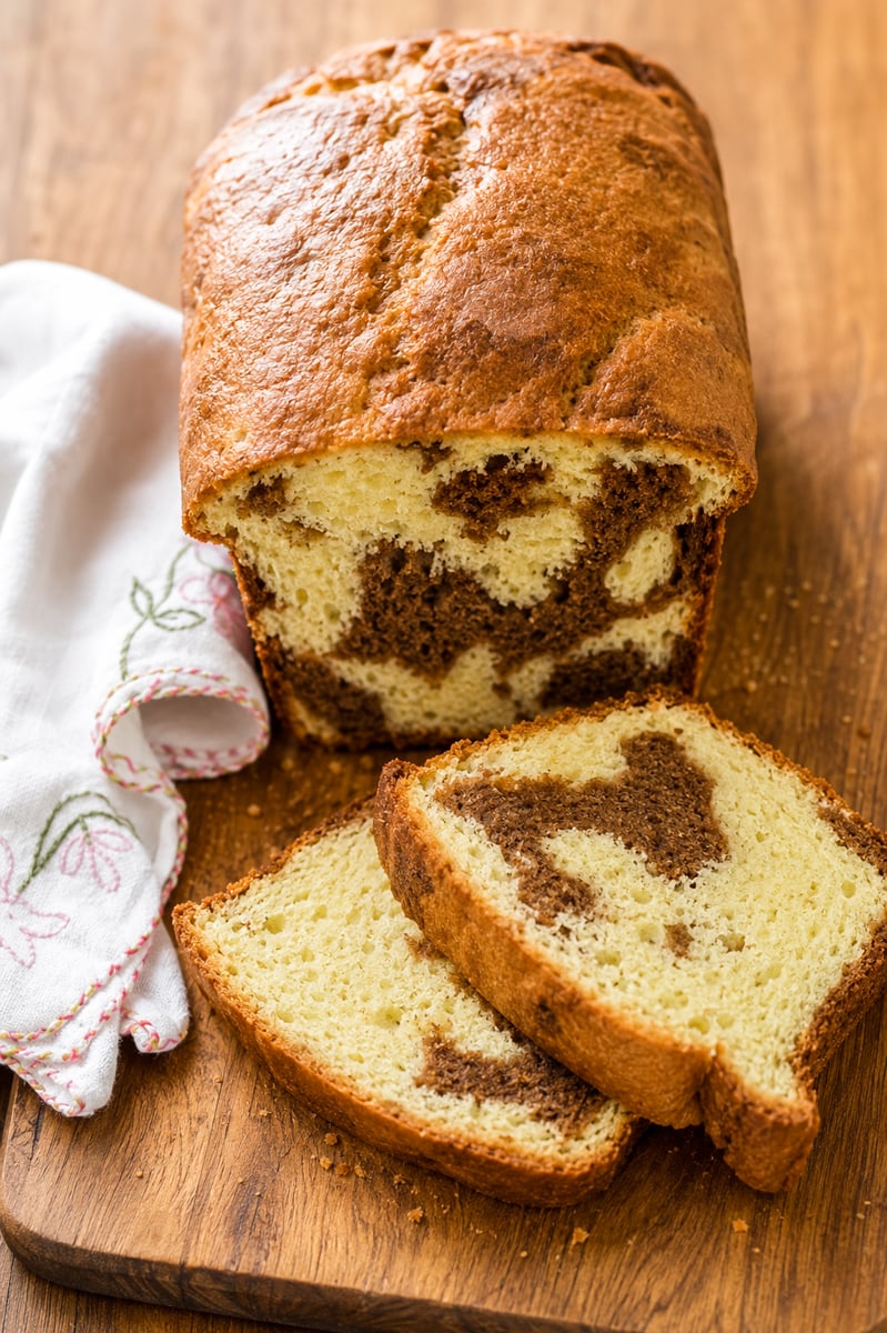 A loaf of marble cake with a golden-brown crust sits on a wooden cutting board. Two slices are cut, showing a swirled chocolate and vanilla pattern. A white cloth with embroidered flowers is beside the bread.