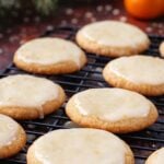 Glazed Lemon Cookies with a glossy glaze sit on a cooling rack. In the background, blurred holiday lights, pine branches, and an orange add festive atmosphere.