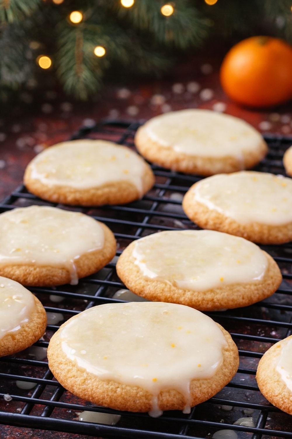 Glazed Lemon Cookies with a glossy glaze sit on a cooling rack. In the background, blurred holiday lights, pine branches, and an orange add festive atmosphere.