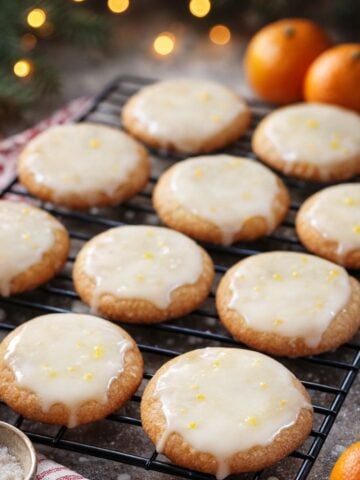 A cooling rack holds round Glazed Lemon Cookies topped with glossy icing and a sprinkle of zest. Oranges and festive lights are blurry in the background, creating a warm, holiday atmosphere.