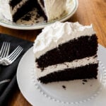 A slice of two-layer chocolate cake with white frosting is served on a white plate, with the rest of the cake in the background on a floral platter. Two forks and a napkin are beside the plate on a wooden table.