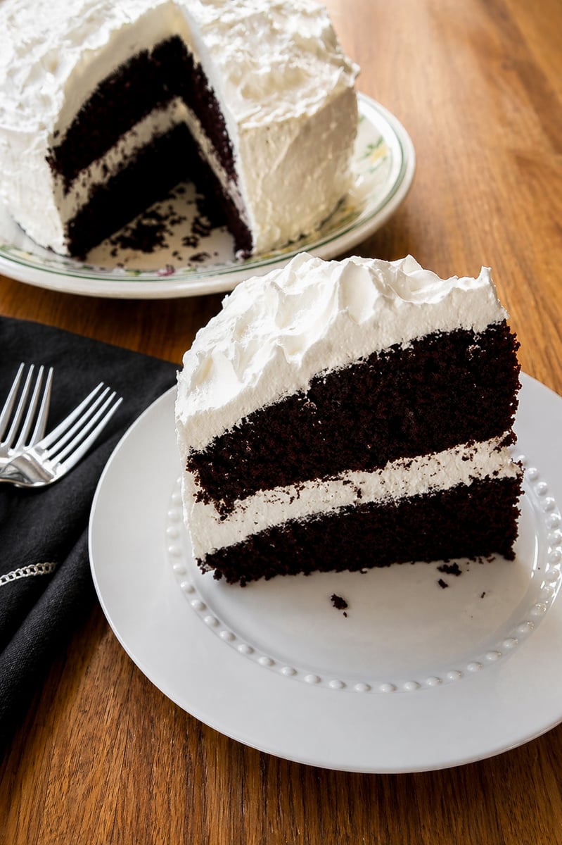 A slice of two-layer chocolate cake with white frosting is served on a white plate, with the rest of the cake in the background on a floral platter. Two forks and a napkin are beside the plate on a wooden table.