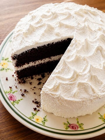 A round chocolate layer cake with white, textured frosting on a floral-patterned plate. A large slice has been cut, showing the dark cake and white filling inside.
