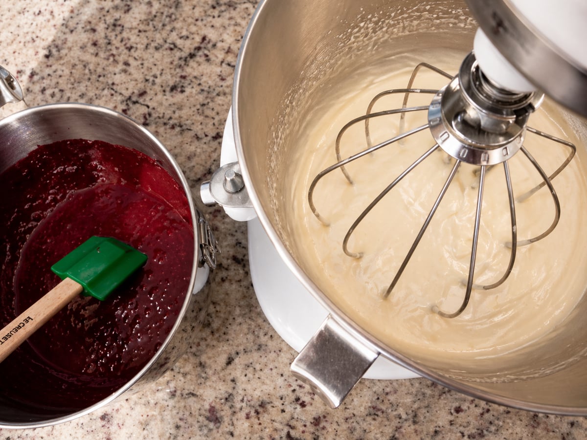 A stand mixer bowl with light cake batter and a metal bowl with red batter and a green spatula sit on a speckled kitchen countertop, ready for baking.