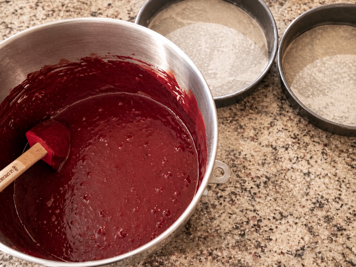 A metal mixing bowl with red cake batter and a spatula sits on a granite countertop next to two round cake pans lined with parchment paper, ready for baking.