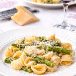 A plate of pasta with asparagus and grated cheese is served on a white tablecloth, with a wedge of cheese, a grater, and a glass of red wine in the background.