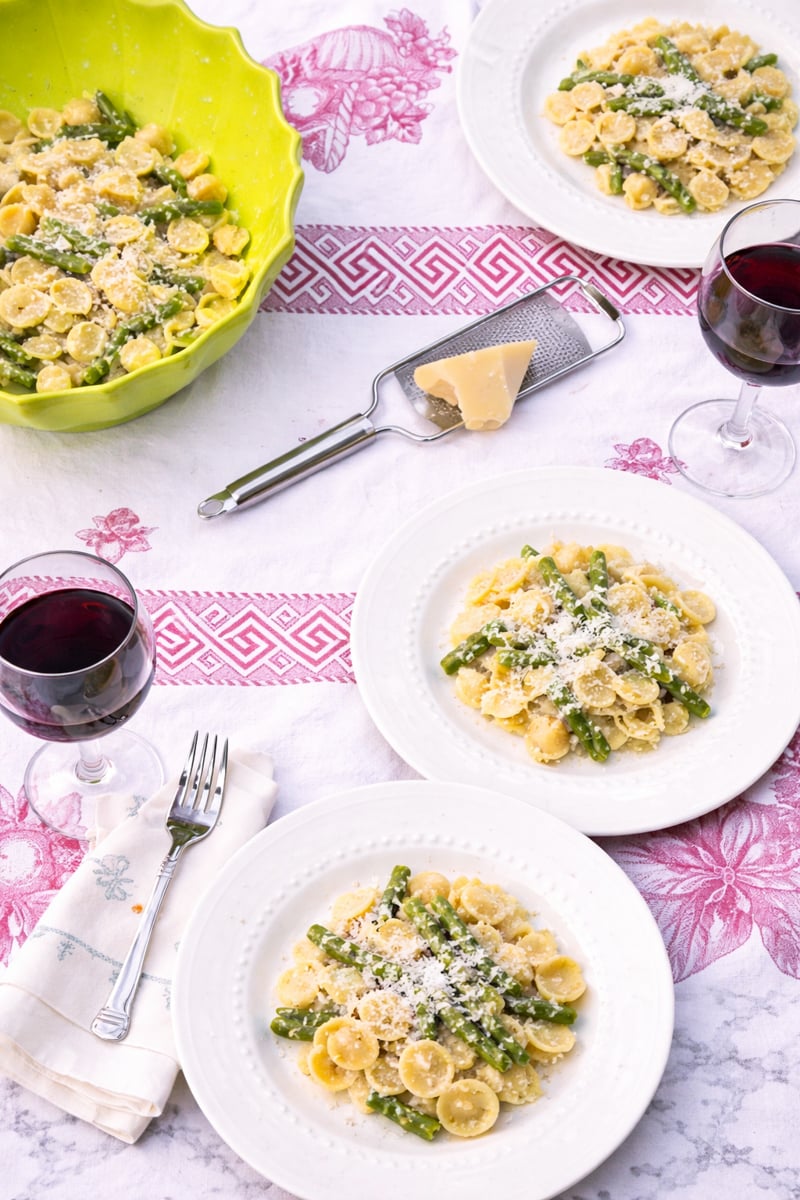 Three plates of pasta with asparagus and grated cheese are set on a table with two glasses of red wine, a bowl of more pasta, a cheese grater, a block of cheese, and a napkin with a fork.