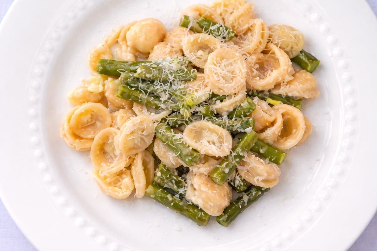 A white plate of orecchiette pasta mixed with pieces of asparagus and topped with grated Parmesan cheese.