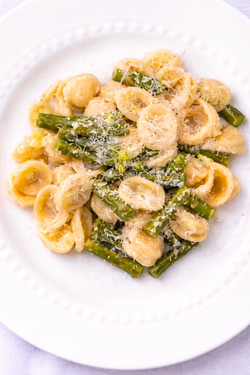 A white plate with orecchiette pasta, asparagus pieces, and a light creamy sauce, topped with grated cheese.