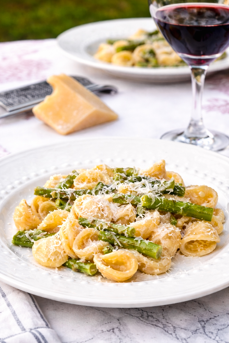 A plate of pasta with asparagus and grated cheese is served on a white tablecloth, with a wedge of cheese, a grater, and a glass of red wine in the background.