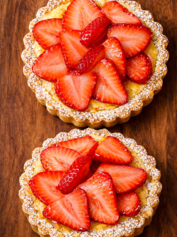 Two small strawberry and ricotta tarts topped with fresh, sliced strawberries and dusted with powdered sugar, placed on a wooden surface.
