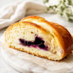A sliced loaf of braided bread with a golden crust and a blueberry filling, displayed on a marble board with a white cloth and blurred greenery in the background.