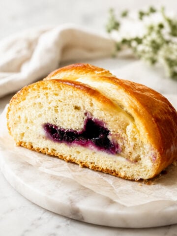 A sliced loaf of braided bread with a golden crust and a blueberry filling, displayed on a marble board with a white cloth and blurred greenery in the background.