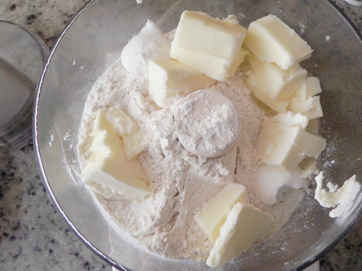 Chunks of butter and flour are placed in a food processor bowl, ready to be mixed for a homemade Sweet Cherry Pie, on a speckled kitchen countertop.