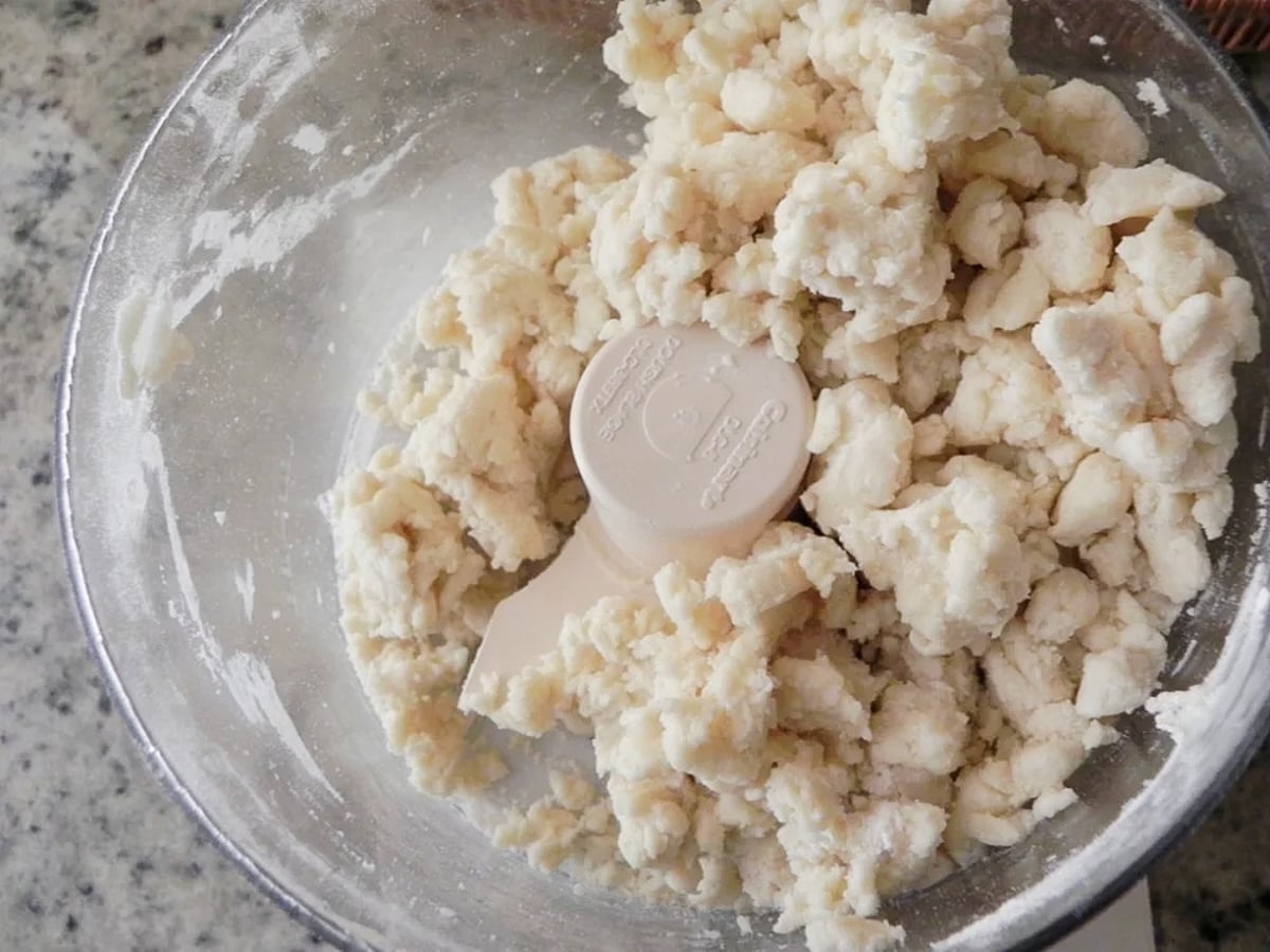 Roughly mixed pie dough for Sweet Cherry Pie in a food processor bowl, with small clumps of flour and butter visible, sitting on a speckled countertop.