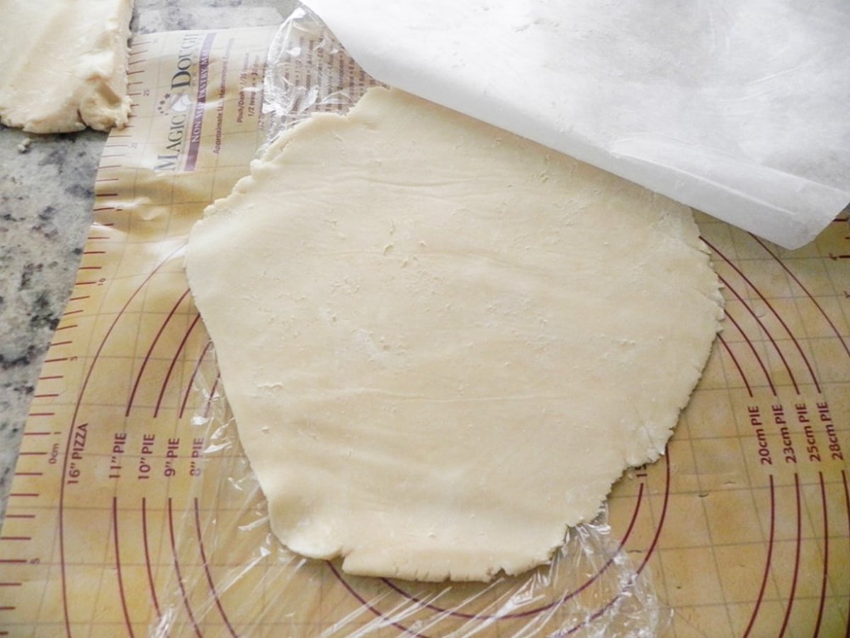 Rolled-out dough for a Sweet Cherry Pie rests on plastic wrap and a pie mat with measurement circles and markings, partially covered by parchment paper, ready for baking or shaping.