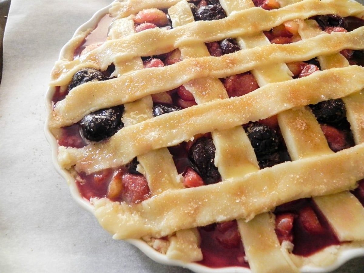 A close-up of an unbaked Sweet Cherry Pie with a lattice crust, showing a mix of red and dark fruit filling beneath golden dough strips sprinkled with sugar. The pie is in a white dish on parchment paper.