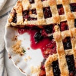 A partially eaten Sweet Cherry Pie with a lattice top sits on a white dish, vibrant red and purple filling peeking through and crumbs scattered about. A striped cloth lies nearby on the marble surface.
