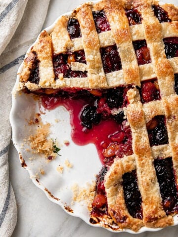 A partially eaten Sweet Cherry Pie with a lattice top sits on a white dish, vibrant red and purple filling peeking through and crumbs scattered about. A striped cloth lies nearby on the marble surface.