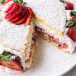 A Summer Strawberry Cake, this round sponge is layered with whipped cream and sliced strawberries, topped with powdered sugar and fresh strawberry slices. One large slice has been cut and slightly removed from the cake.
