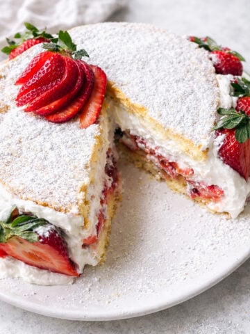 A Summer Strawberry Cake, this round sponge is layered with whipped cream and sliced strawberries, topped with powdered sugar and fresh strawberry slices. One large slice has been cut and slightly removed from the cake.