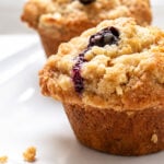 Two blueberry muffins with crumbly streusel topping sit on a white plate. A few crumbs are scattered nearby, and the background is softly blurred.