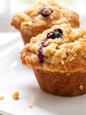 Two blueberry muffins with crumbly streusel topping sit on a white plate. A few crumbs are scattered nearby, and the background is softly blurred.