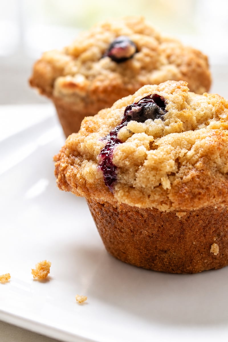 Two blueberry muffins with crumbly streusel topping sit on a white plate. A few crumbs are scattered nearby, and the background is softly blurred.