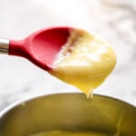 A red silicone spatula holding thick, creamy yellow pastry cream above a metal bowl filled with more custard, set on a light-colored surface.