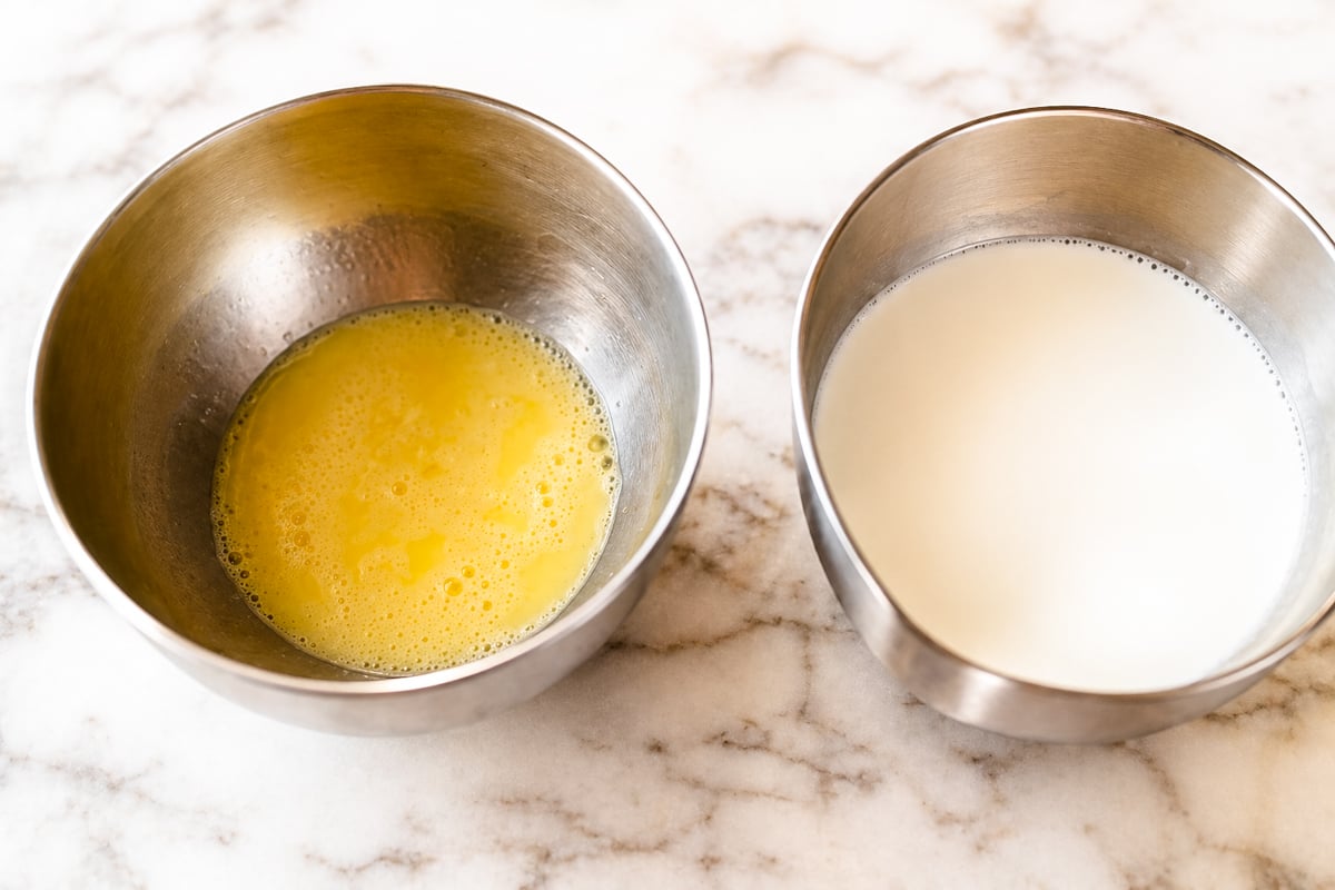 Two metal bowls on a marble surface: one holds beaten eggs, while the other has milk-essential ingredients for making silky pastry cream.
