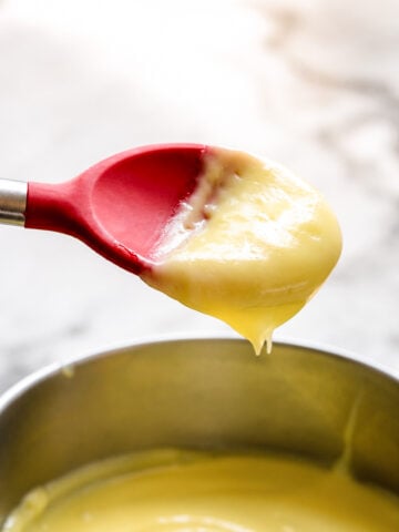 A red silicone spatula holding thick, creamy yellow pastry cream above a metal bowl filled with more custard, set on a light-colored surface.
