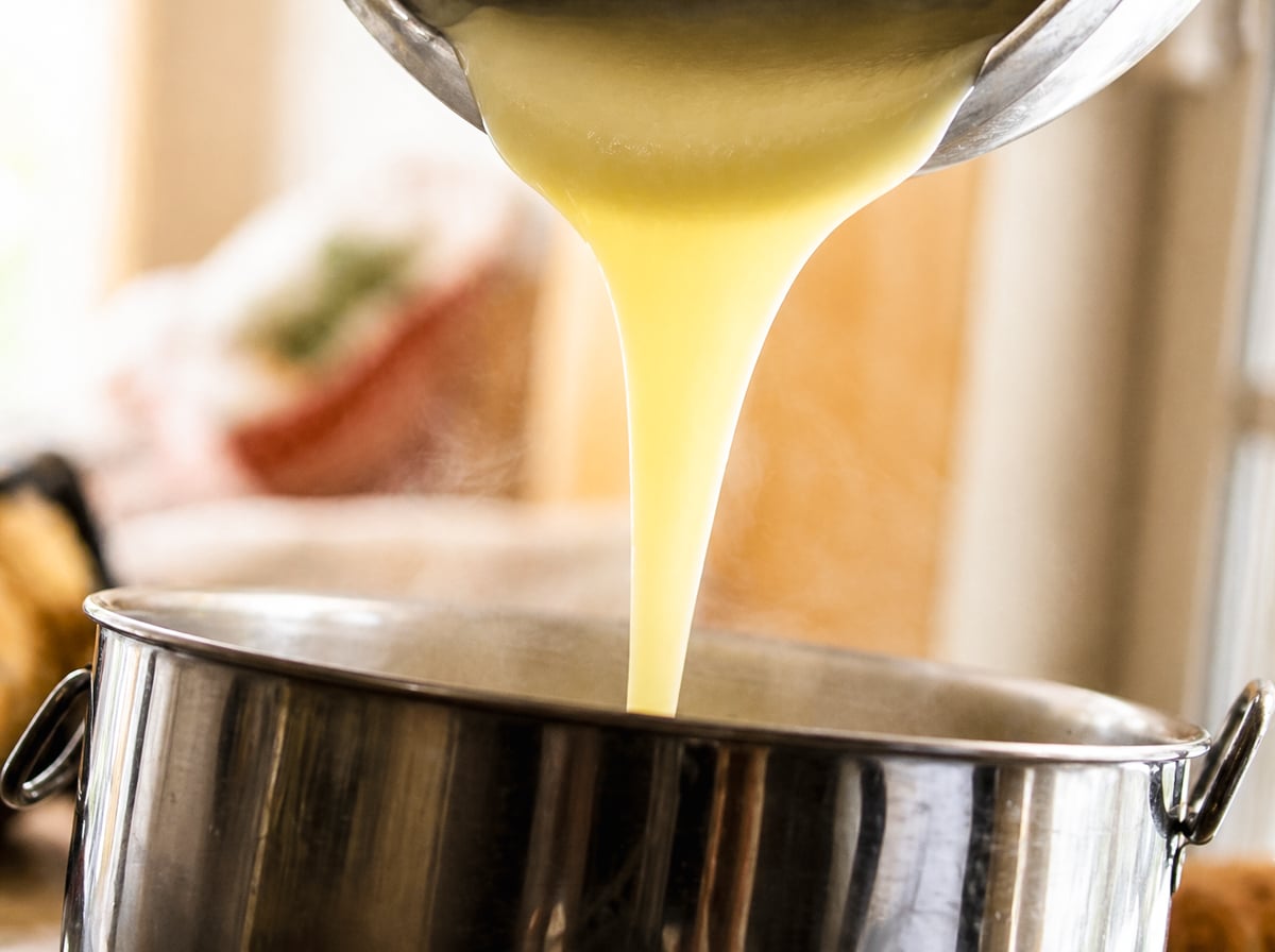A thick, creamy pastry cream is being poured from a metal bowl into a large stainless steel pot, with steam rising from the pot.