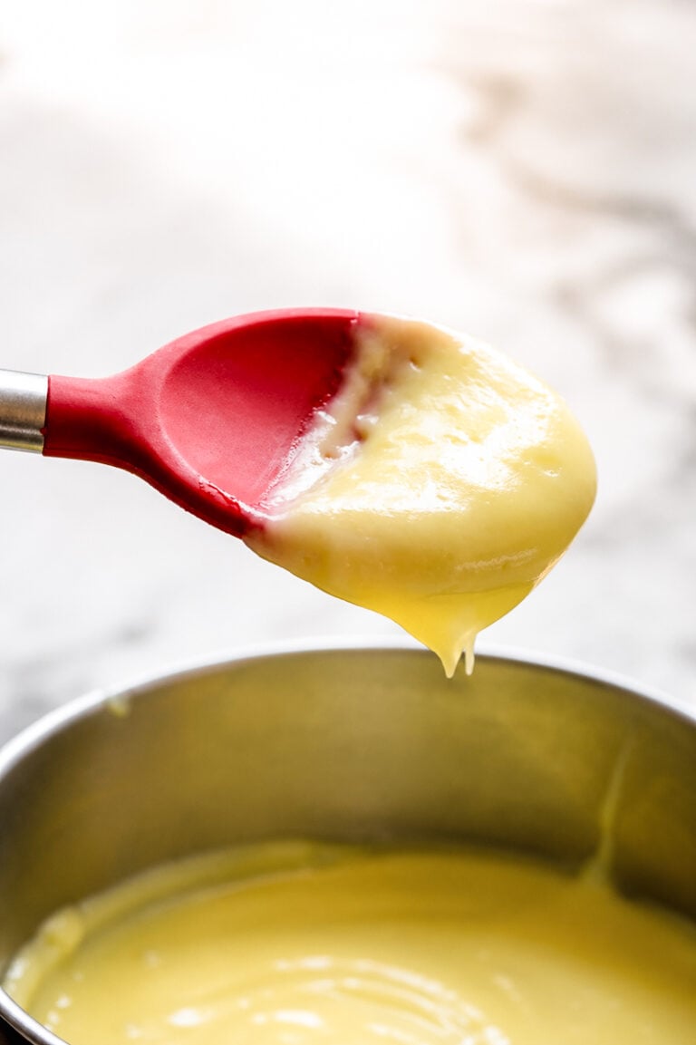 A red silicone spatula holding thick, creamy yellow pastry cream above a metal bowl filled with more custard, set on a light-colored surface.