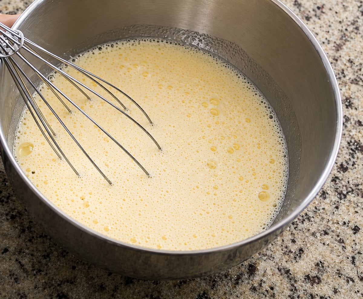 A metal whisk sits in a stainless steel bowl filled with light yellow, frothy pastry cream mixture on a speckled countertop.