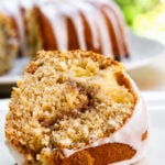 A close-up of a Peach Cinnamon Swirl Bundt Cake slice reveals a swirl of cinnamon and apple chunks, topped with a thin white glaze. The rest of the cake sits in the background, slightly out of focus.