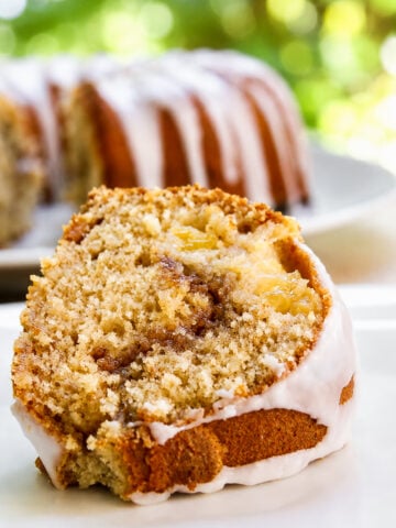 A close-up of a Peach Cinnamon Swirl Bundt Cake slice reveals a swirl of cinnamon and apple chunks, topped with a thin white glaze. The rest of the cake sits in the background, slightly out of focus.