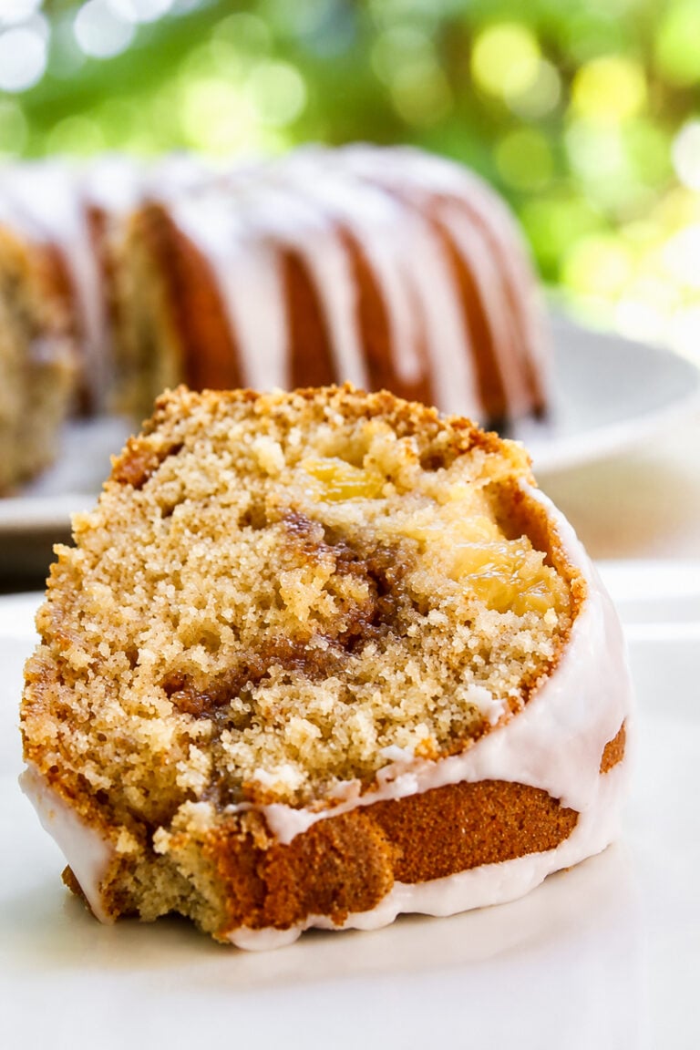 A close-up of a Peach Cinnamon Swirl Bundt Cake slice reveals a swirl of cinnamon and apple chunks, topped with a thin white glaze. The rest of the cake sits in the background, slightly out of focus.