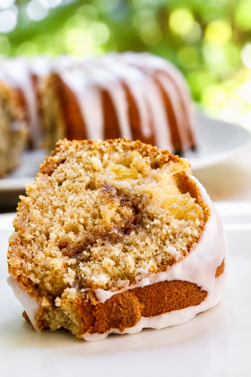 A close-up of a Peach Cinnamon Swirl Bundt Cake slice reveals a swirl of cinnamon and apple chunks, topped with a thin white glaze. The rest of the cake sits in the background, slightly out of focus.