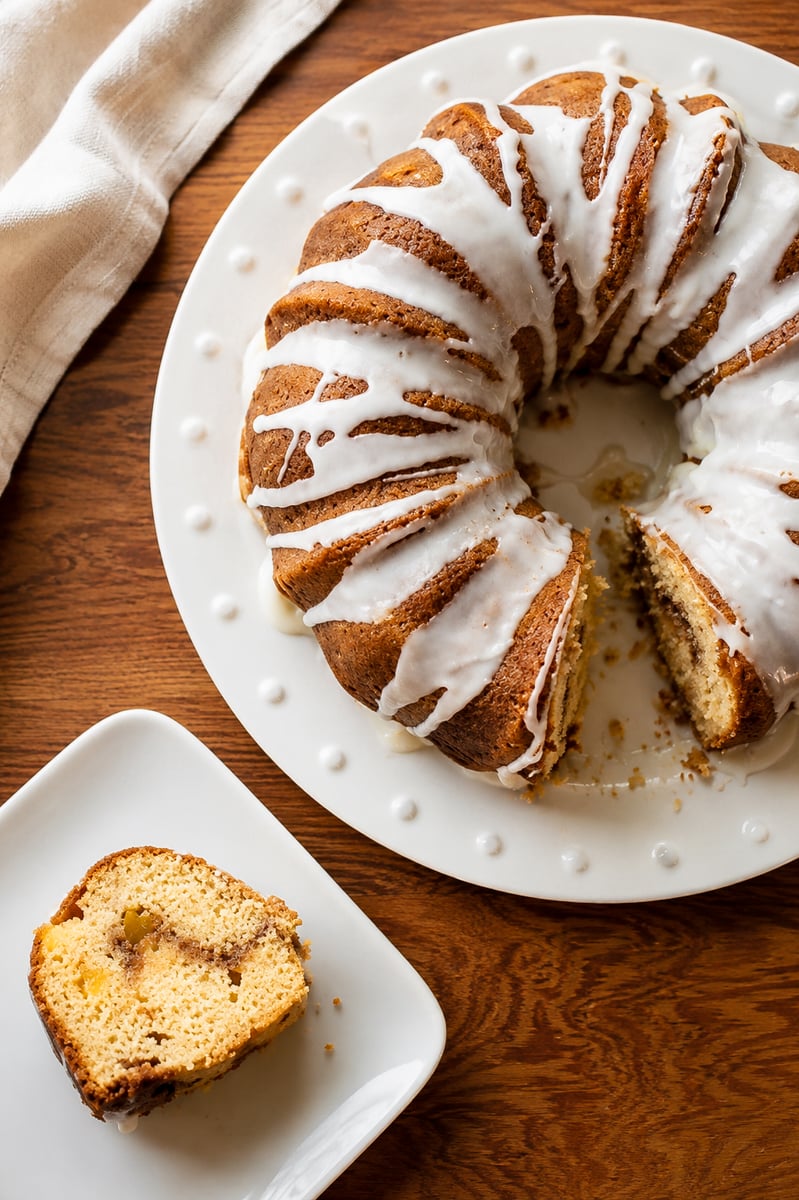 A Peach Cinnamon Swirl Bundt Cake with white icing sits on a round white plate; a slice is served on a square dish nearby. The cake features cinnamon swirls and rests on a wooden table with a cloth napkin.